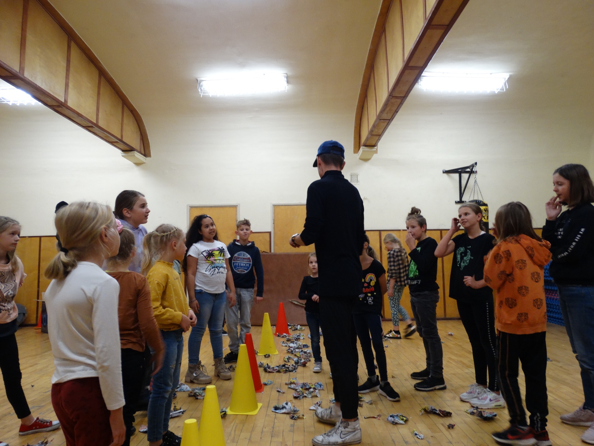 Animator w Łodzi prowadzi grę z dziećmi w sali gimnastycznej, wykorzystując kolorowe pachołki i porozrzucane na podłodze papierki.