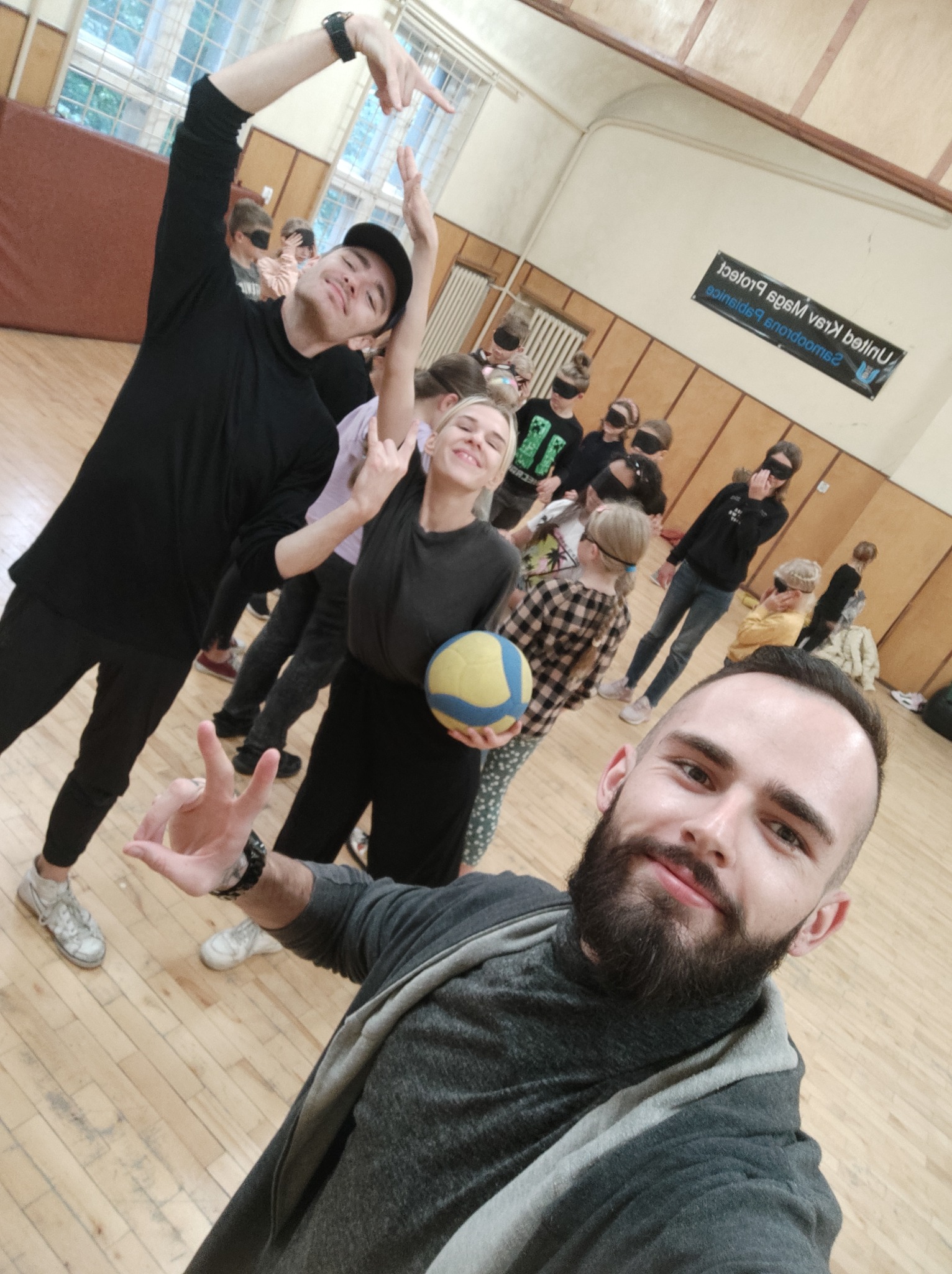 Grupa dzieci z zasłoniętymi oczami uczestniczy w zajęciach w sali gimnastycznej, prowadzonych przez animatorów, jeden z animatorów robi selfie.