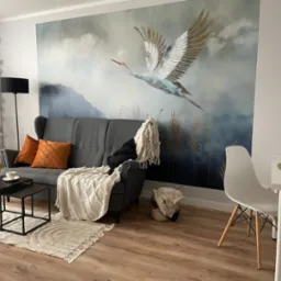 Szary salon z fototapetą przedstawiającą żurawia w locie, sofa z pomarańczowymi poduszkami i białym pledem, stolik kawowy z czarnymi nogami, dywan z frędzlami i białe krzesła przy stole.