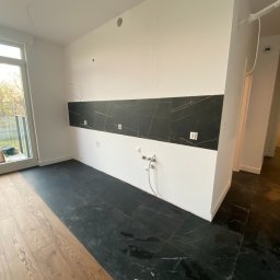 MercBud Sp z o o - Częściowo wykończona kuchnia z czarną marmurową podłogą i pasem płytek nad blatem, widoczne instalacje elektryczne i wodne, podłoga częściowo wyłożona drewnianymi panelami.