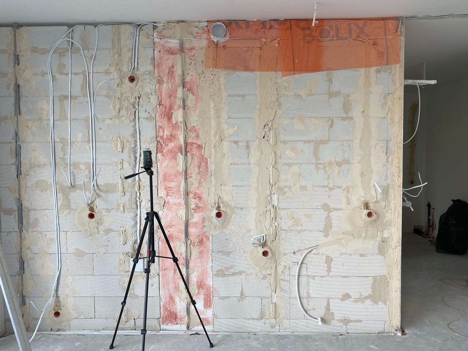 Ściana z białych bloczków z widocznymi przewodami elektrycznymi, częściowo pokryta szpachlą i zabezpieczona folią, z laserowym niwelatorem na statywie.