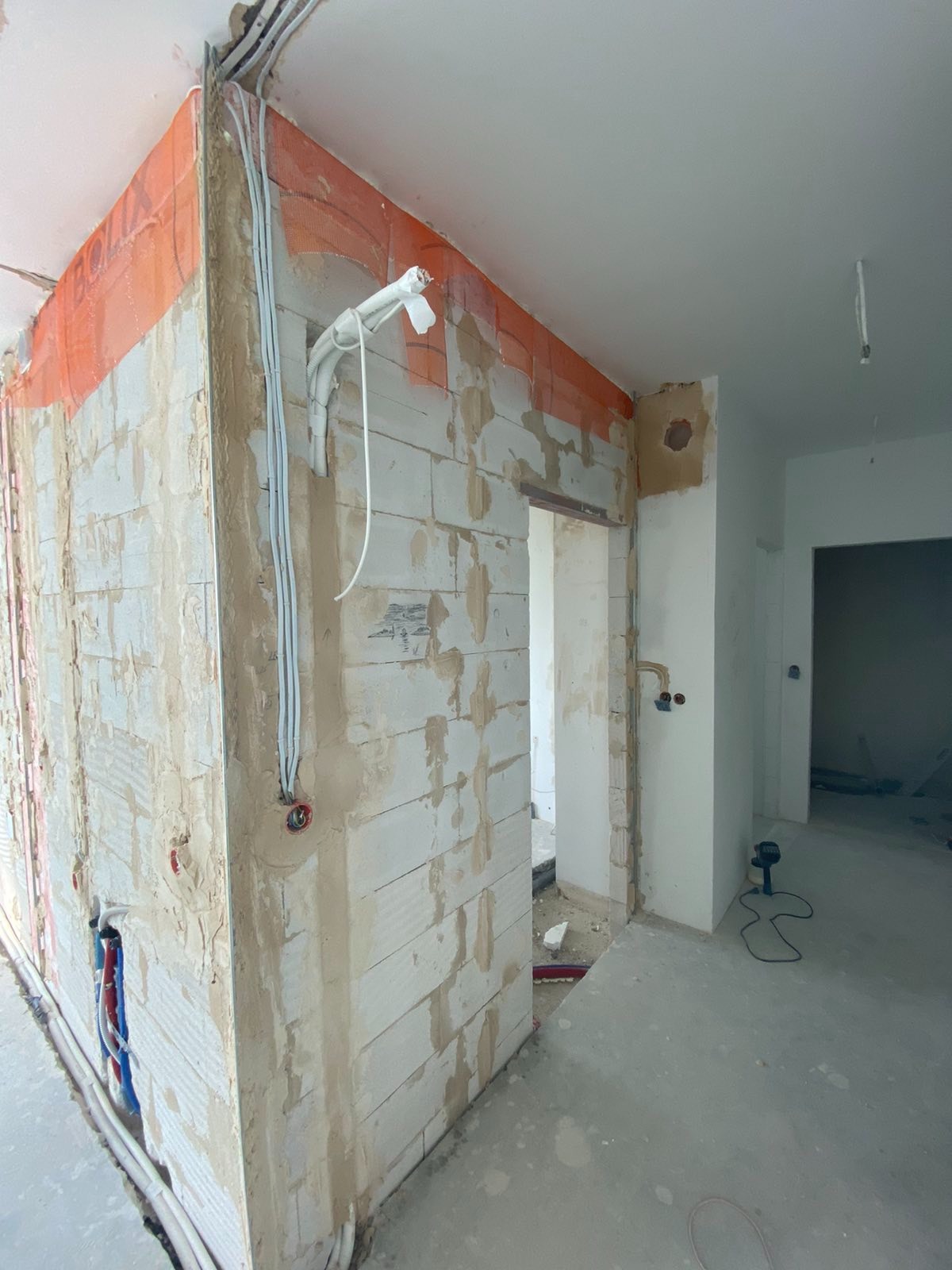 Wnętrze w trakcie remontu: ściany z białych bloczków, częściowo pokryte zaprawą, widoczne przewody elektryczne i rury, zabezpieczone narożniki pomarańczową siatką, surowy beton na podłodze.
