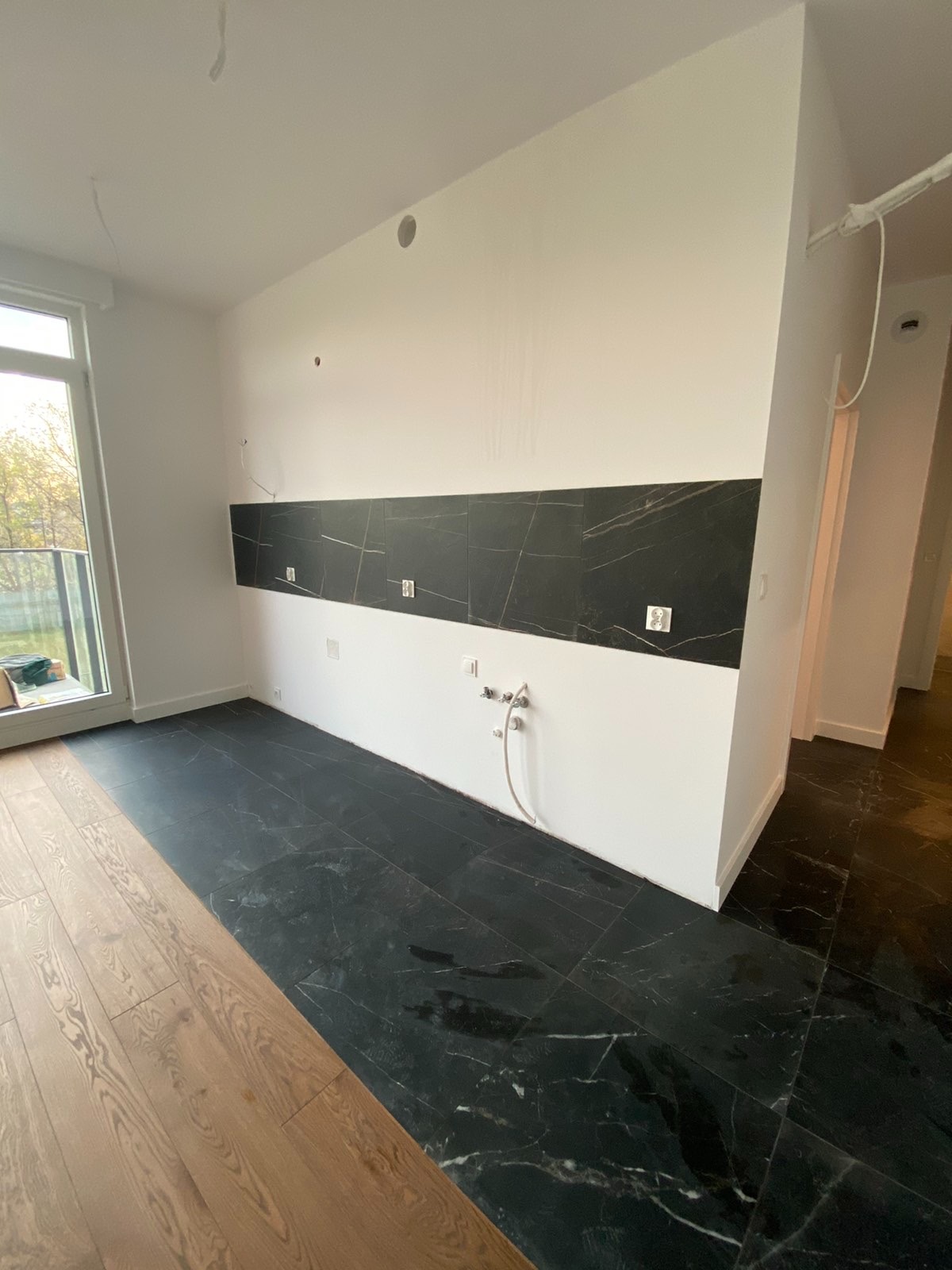 Częściowo wykończona kuchnia z czarną marmurową podłogą i pasem płytek nad blatem, widoczne instalacje elektryczne i wodne, podłoga częściowo wyłożona drewnianymi panelami.