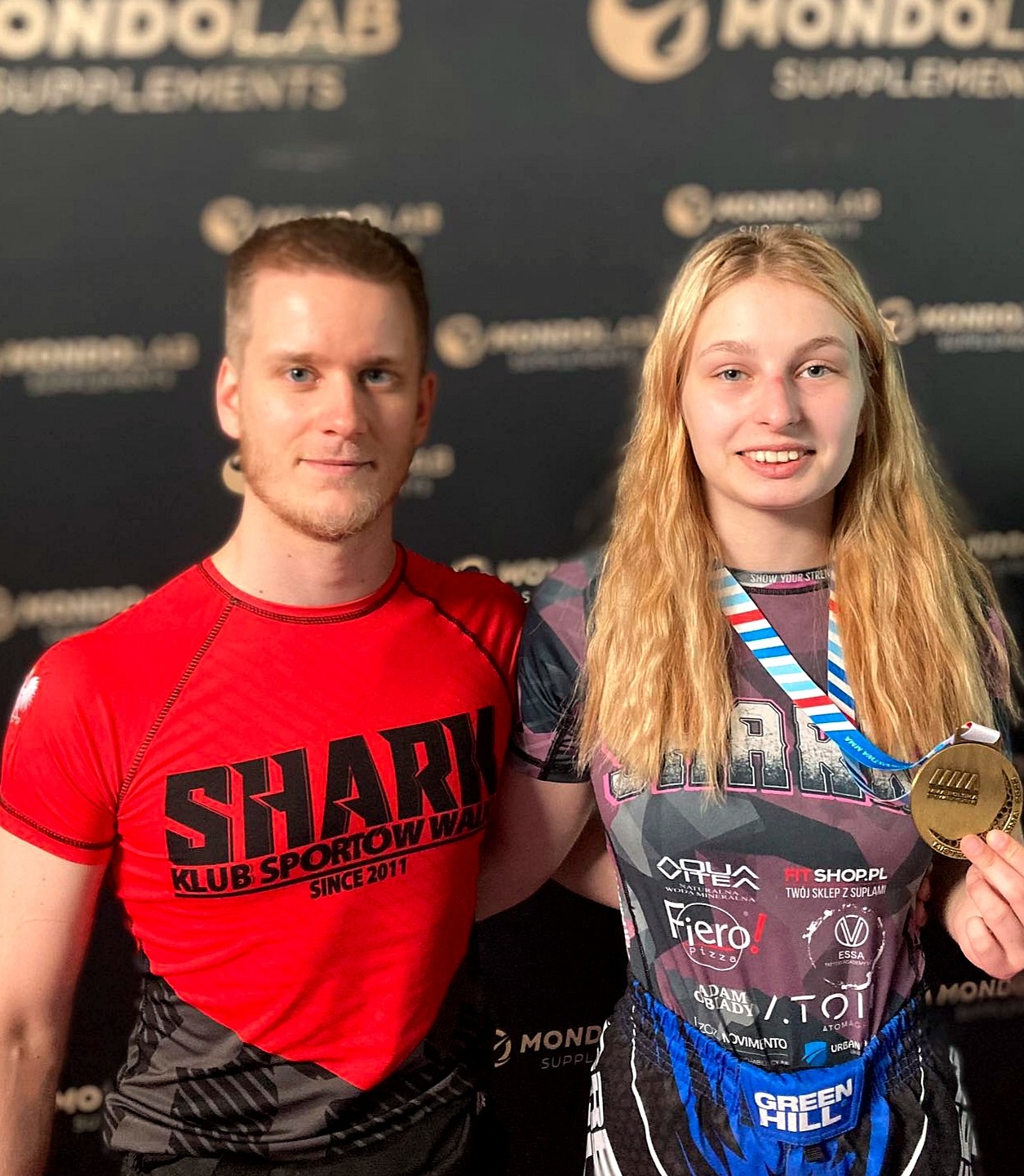 Młoda zawodniczka MMA, Anastasiia Holovko, pozuje z trenerem po zdobyciu złotego medalu na turnieju MMA Polska 8, trzymając medal w dłoni, oboje ubrani w odzież sportową z logo klubu.