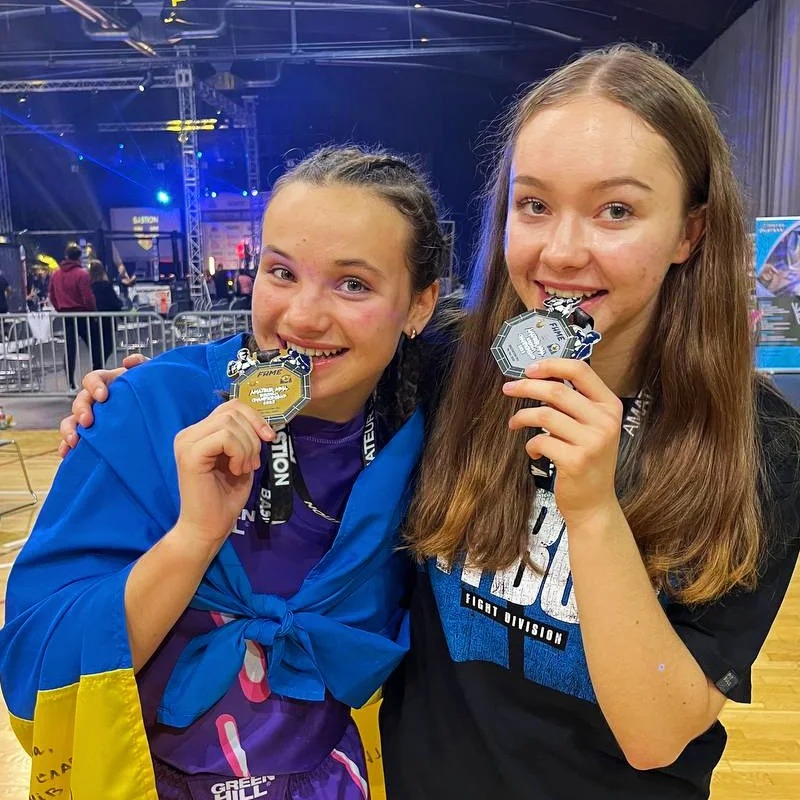 Dwie młode zawodniczki MMA, trzymające medale Mistrzostw Europy 2023, uśmiechają się do kamery; jedna owinięta flagą Ukrainy, druga w czarnej koszulce z napisem 'FIGHT DIVISION'.