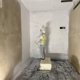 Malowanie natryskowe ścian w pomieszczeniu. Pracownik w kombinezonie ochronnym maluje ścianę na biało. Podłoga zabezpieczona folią. Częściowo pomalowana ściana.