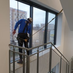 Paweł Przybyszewski-specjalistyczne doczyszczanie - Pracownik w uprzęży alpinistycznej, z widocznym sprzętem, czyści okno od zewnątrz w wysokim budynku, widok z klatki schodowej.