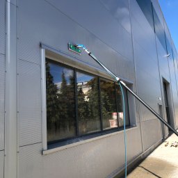 Paweł Przybyszewski-specjalistyczne doczyszczanie - Mycie okna na elewacji budynku przemysłowego za pomocą teleskopowej lancy z szczotką, widoczne krople wody i odbicie drzew w oknie.