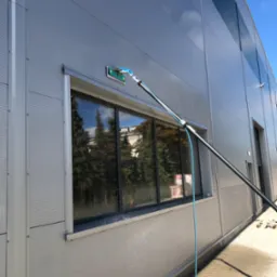 Mycie okna na elewacji budynku przemysłowego za pomocą teleskopowej lancy z szczotką, widoczne krople wody i odbicie drzew w oknie.