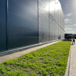 Paweł Przybyszewski-specjalistyczne doczyszczanie - Pracownik w odblaskowej kamizelce czyści elewację przemysłowego budynku za pomocą lancy teleskopowej, widoczne ślady wody na panelach oraz świeża trawa wzdłuż ścieżki.