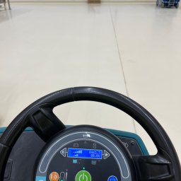 Paweł Przybyszewski-specjalistyczne doczyszczanie - Widok z perspektywy operatora na kontrolki i kierownicę maszyny czyszczącej posadzkę w hali przemysłowej; w tle widoczna paleta i podnośnik koszowy.