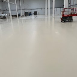 Paweł Przybyszewski-specjalistyczne doczyszczanie - Błyszcząca, świeżo pomalowana podłoga w hali przemysłowej, widoczny podnośnik nożycowy Skyjack, białe ściany i słupy konstrukcyjne, odległe drzwi i elementy wyposażenia.