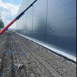 Paweł Przybyszewski-specjalistyczne doczyszczanie - Czyszczenie elewacji przemysłowej za pomocą teleskopowej lancy z dyszą, widoczne zabrudzenia na powierzchni, przygotowany teren wokół budynku z rozłożonym przewodem zasilającym.