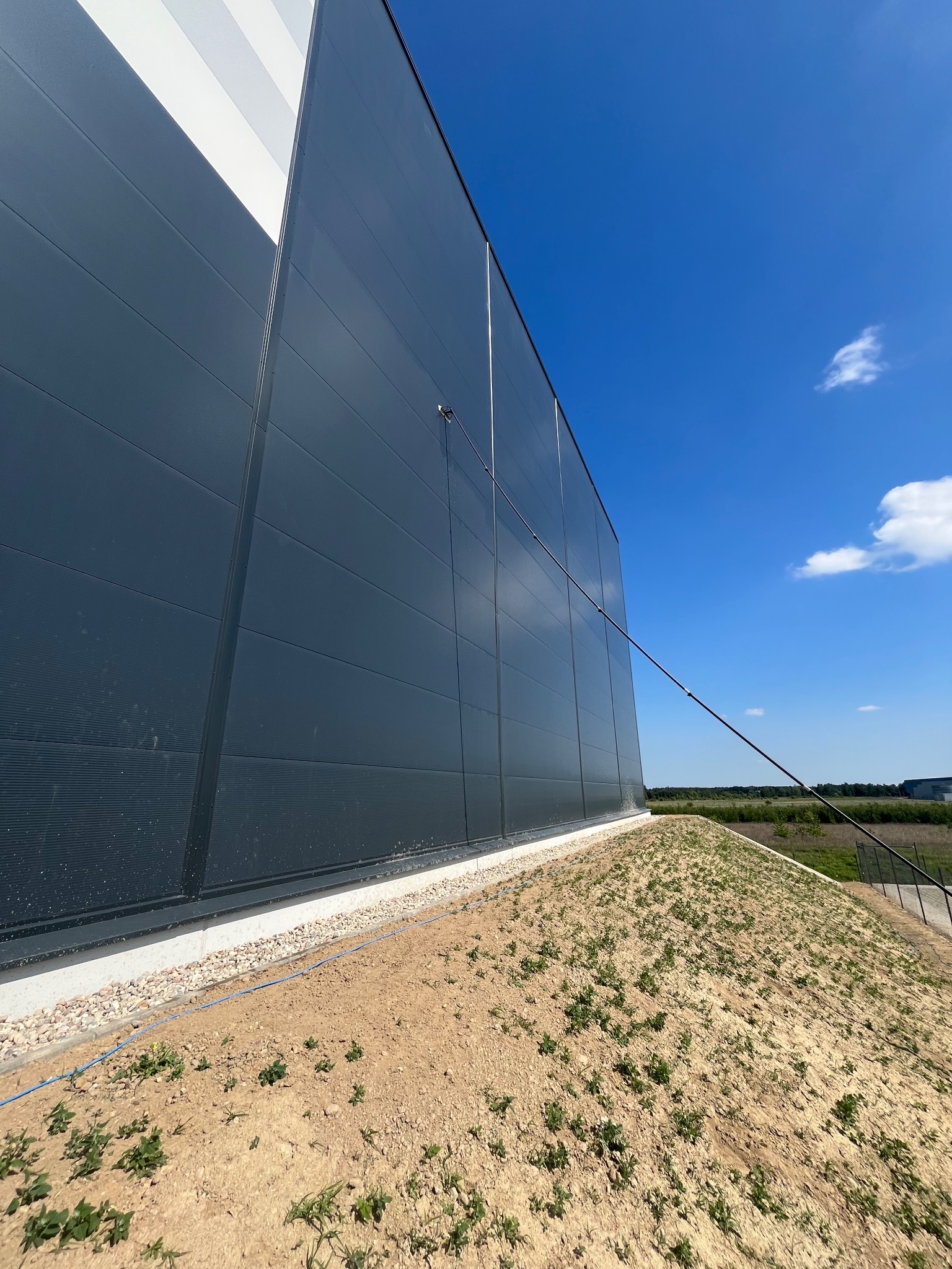 Czyszczenie elewacji przemysłowej z użyciem lancy teleskopowej. Szara, panelowa ściana budynku i błękitne niebo w tle.