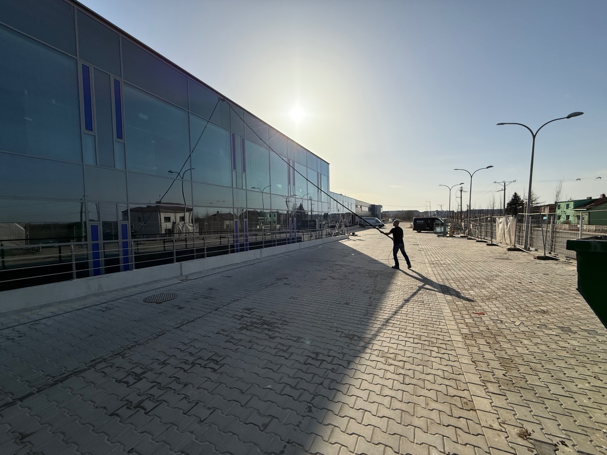 Mycie przeszklonej fasady budynku komercyjnego z użyciem lancy teleskopowej. Osoba w trakcie pracy, widoczne refleksy słońca na szklanych powierzchniach.