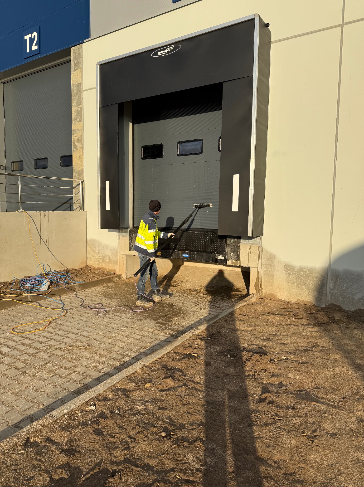 Pracownik w kamizelce czyści bramę przemysłową myjką ciśnieniową. Widoczne ślady czyszczenia, kostka brukowa i zwinięte węże. Brama marki Novoferm.
