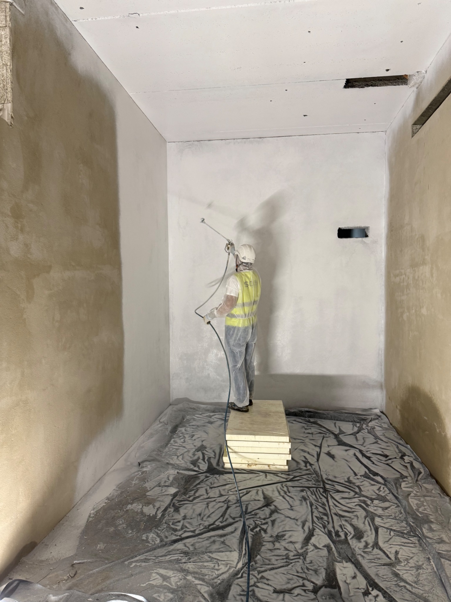 Malowanie natryskowe ścian w pomieszczeniu. Pracownik w kombinezonie ochronnym maluje ścianę na biało. Podłoga zabezpieczona folią. Częściowo pomalowana ściana.