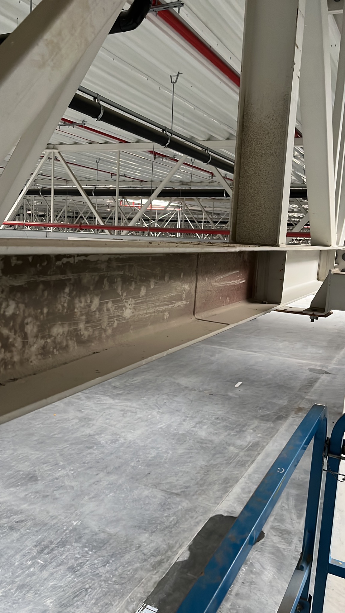 Wnętrze hali przemysłowej z widoczną konstrukcją stalową, rurami pod sufitem i fragmentem niebieskiego podnośnika koszowego w dolnym rogu.