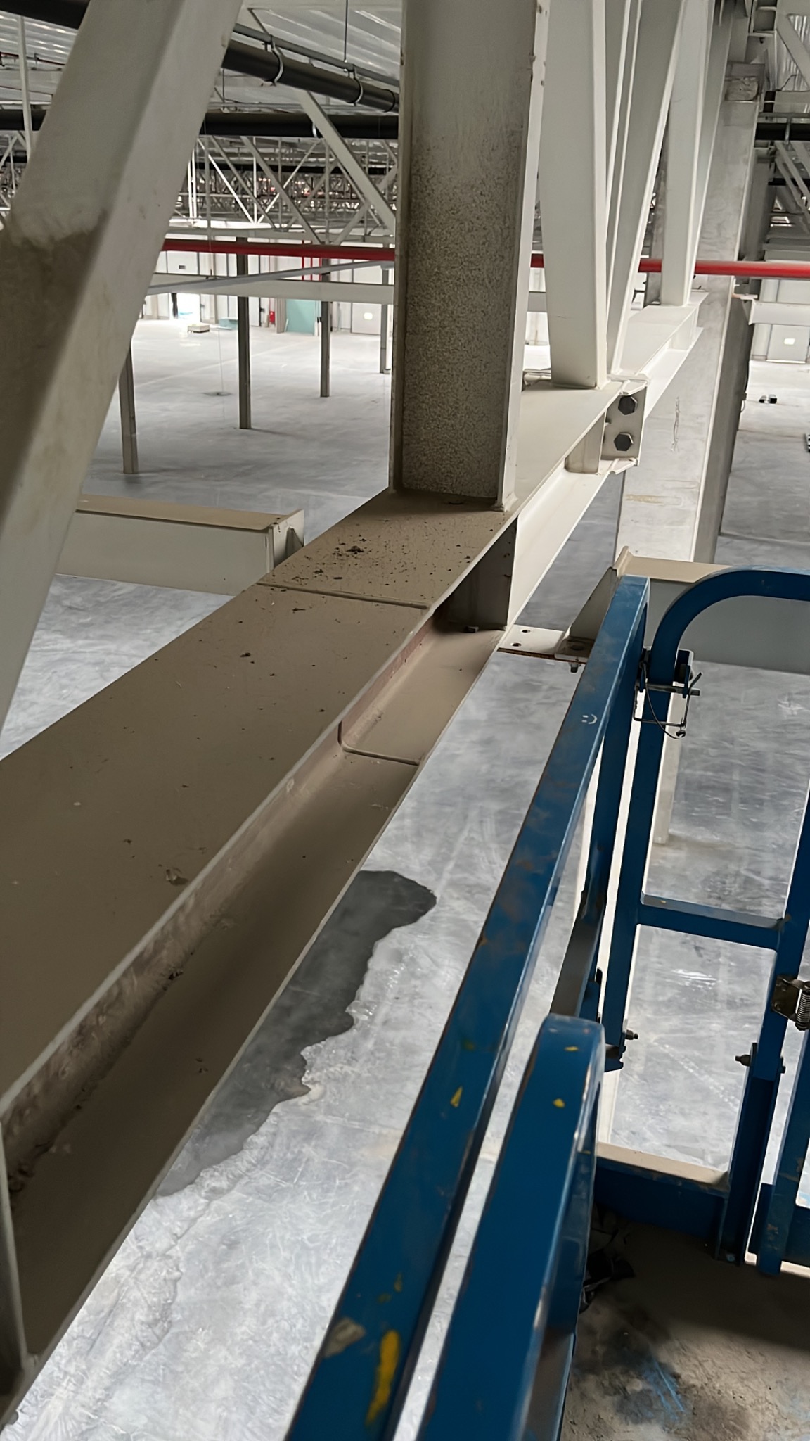 Metalowa konstrukcja hali przemysłowej z widocznym brudem i kurzem, fragment podnośnika koszowego w kolorze niebieskim, szara posadzka z plamami wody.