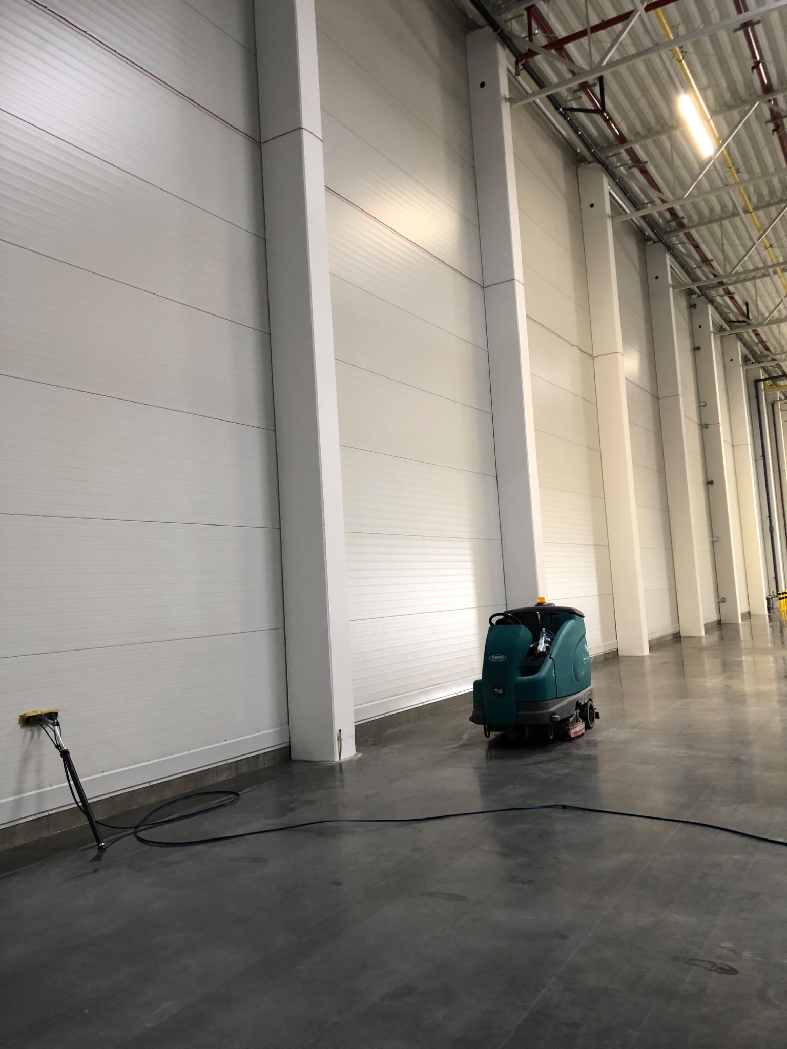 Automat szorujący Tennant T16 na posadzce przemysłowej, widoczny również wąż i szczotka do czyszczenia narożników, oświetlenie LED na suficie.