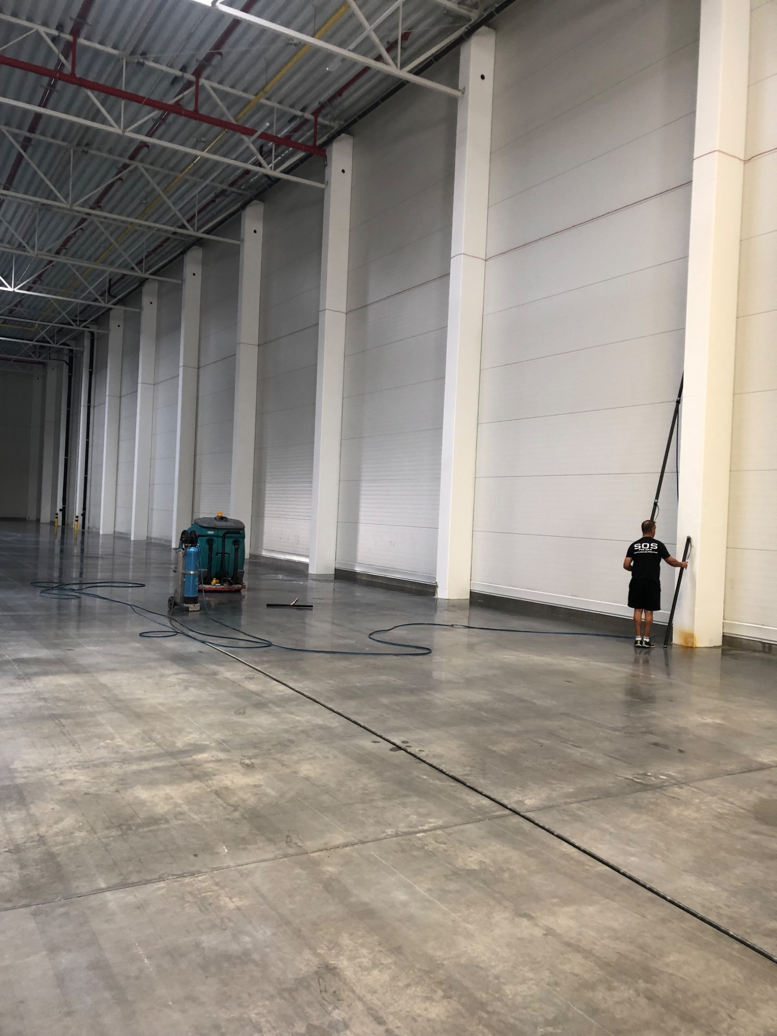 Pracownik w czarnej koszulce z logo czyści wysoką ścianę hali przemysłowej za pomocą długiego kija z padem, obok stoi maszyna czyszcząca z niebieskim zbiornikiem i wężami na posadzce