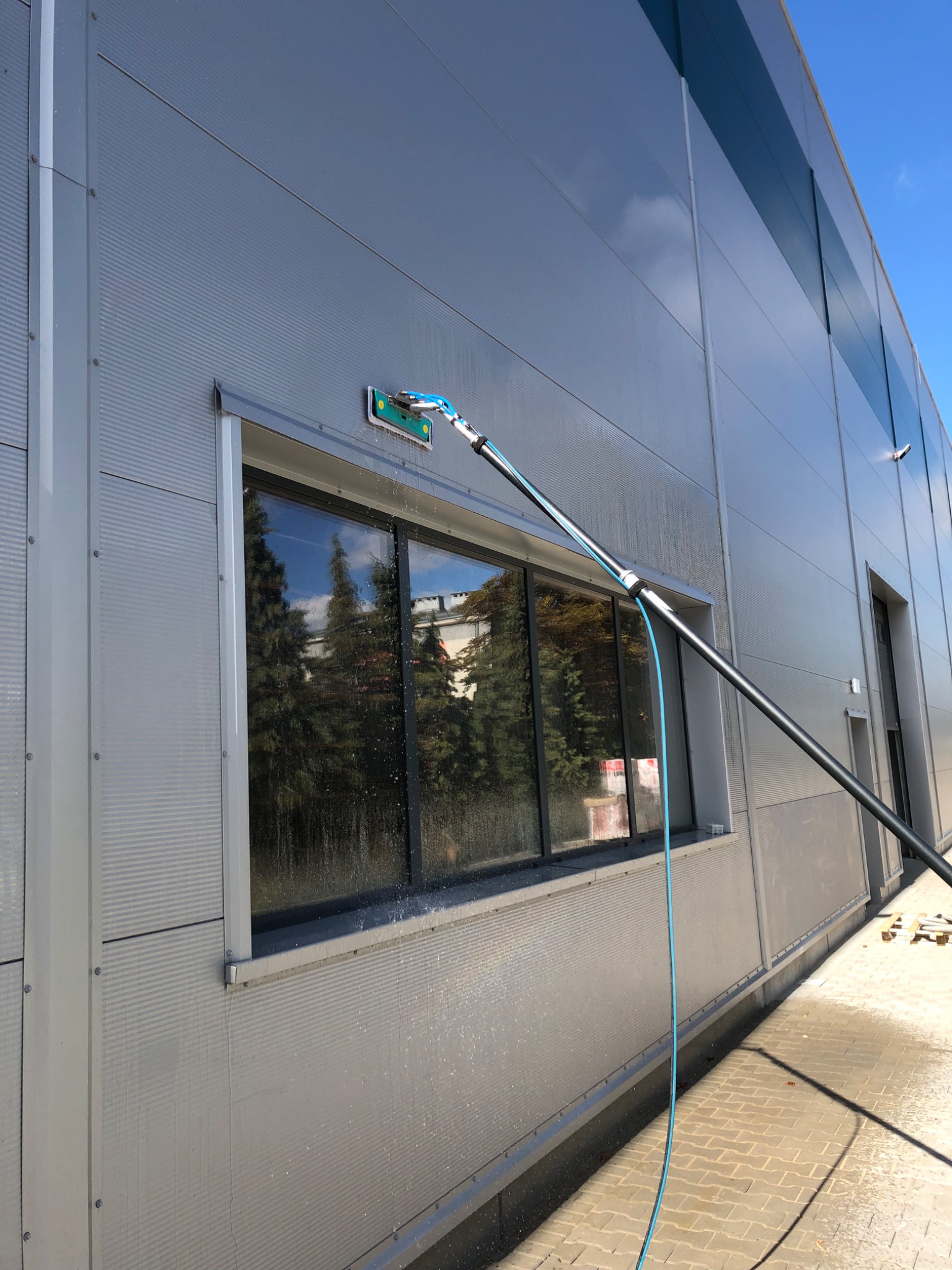 Mycie okna na elewacji budynku przemysłowego za pomocą teleskopowej lancy z szczotką, widoczne krople wody i odbicie drzew w oknie.
