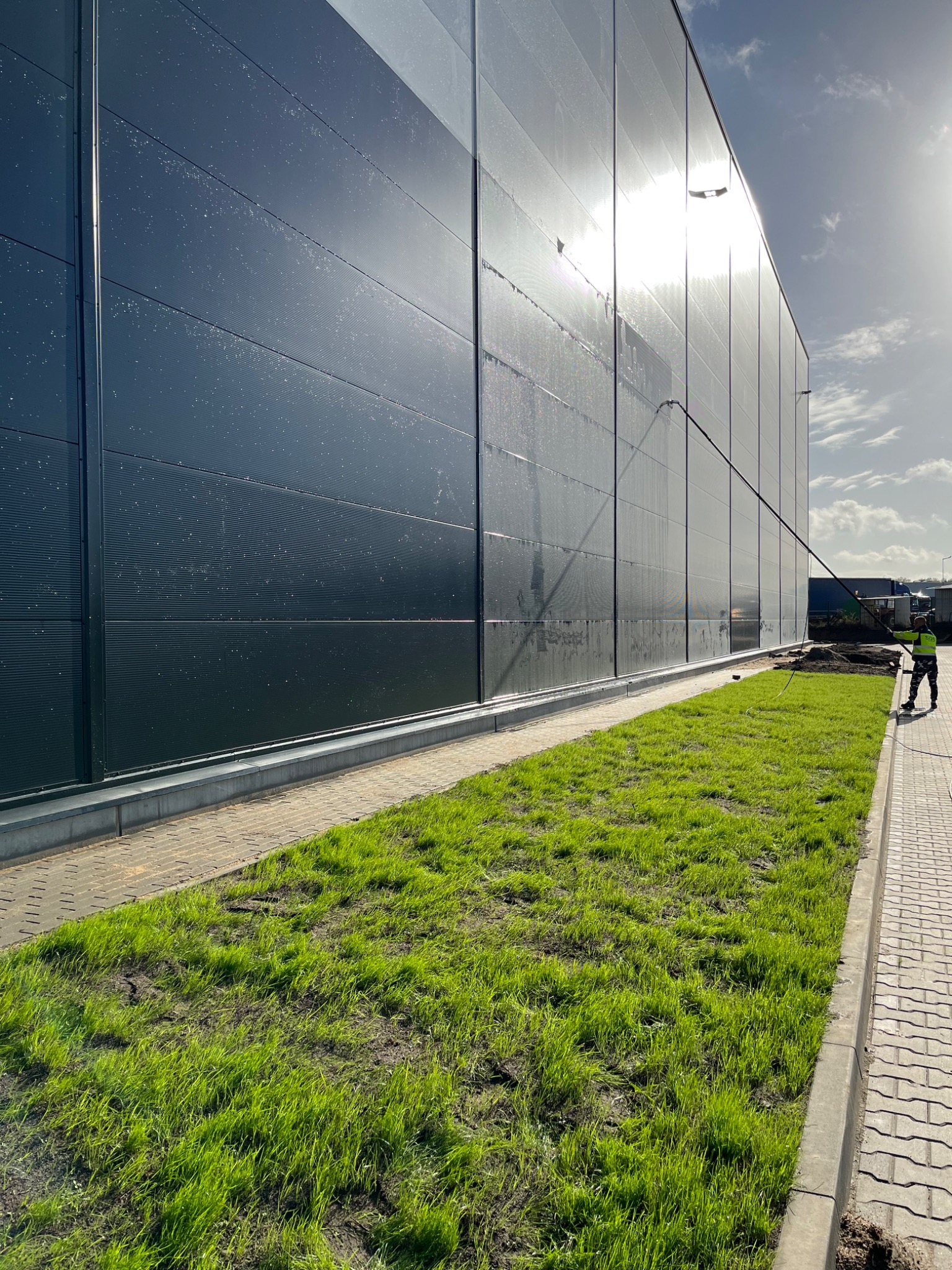Pracownik w odblaskowej kamizelce czyści elewację przemysłowego budynku za pomocą lancy teleskopowej, widoczne ślady wody na panelach oraz świeża trawa wzdłuż ścieżki.