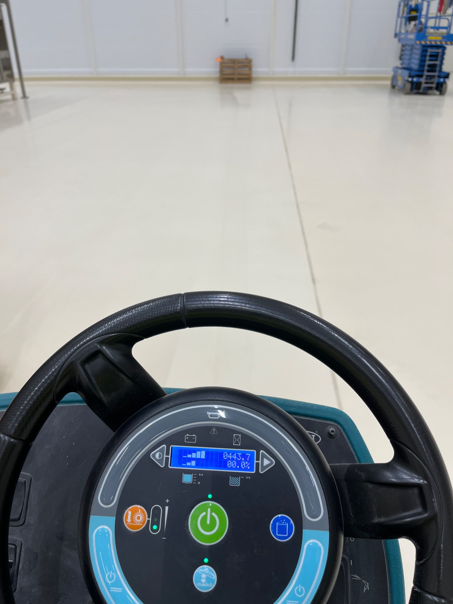 Widok z perspektywy operatora na kontrolki i kierownicę maszyny czyszczącej posadzkę w hali przemysłowej; w tle widoczna paleta i podnośnik koszowy.