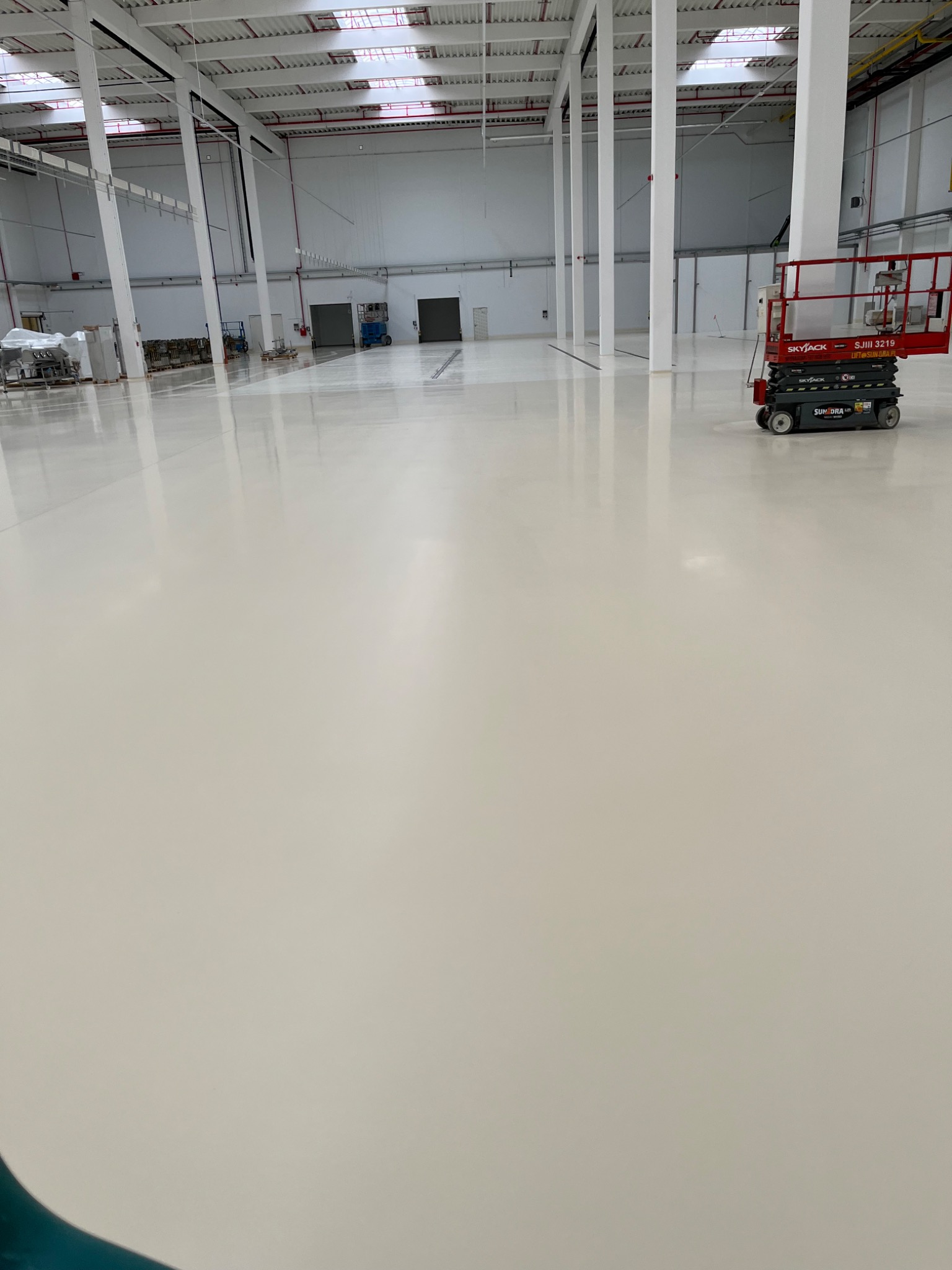 Błyszcząca, świeżo pomalowana podłoga w hali przemysłowej, widoczny podnośnik nożycowy Skyjack, białe ściany i słupy konstrukcyjne, odległe drzwi i elementy wyposażenia.
