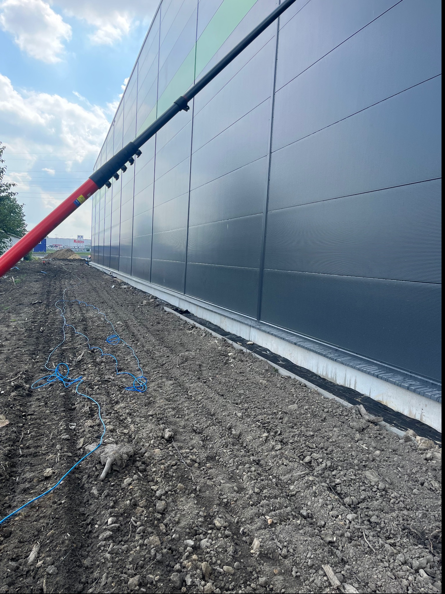 Czyszczenie elewacji przemysłowej za pomocą teleskopowej lancy z dyszą, widoczne zabrudzenia na powierzchni, przygotowany teren wokół budynku z rozłożonym przewodem zasilającym.