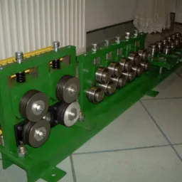 Zielona maszyna do obróbki metalu z szeregiem metalowych rolek i żółtą osłoną, ustawiona na kafelkowanej podłodze, z widocznym grzejnikiem w tle.