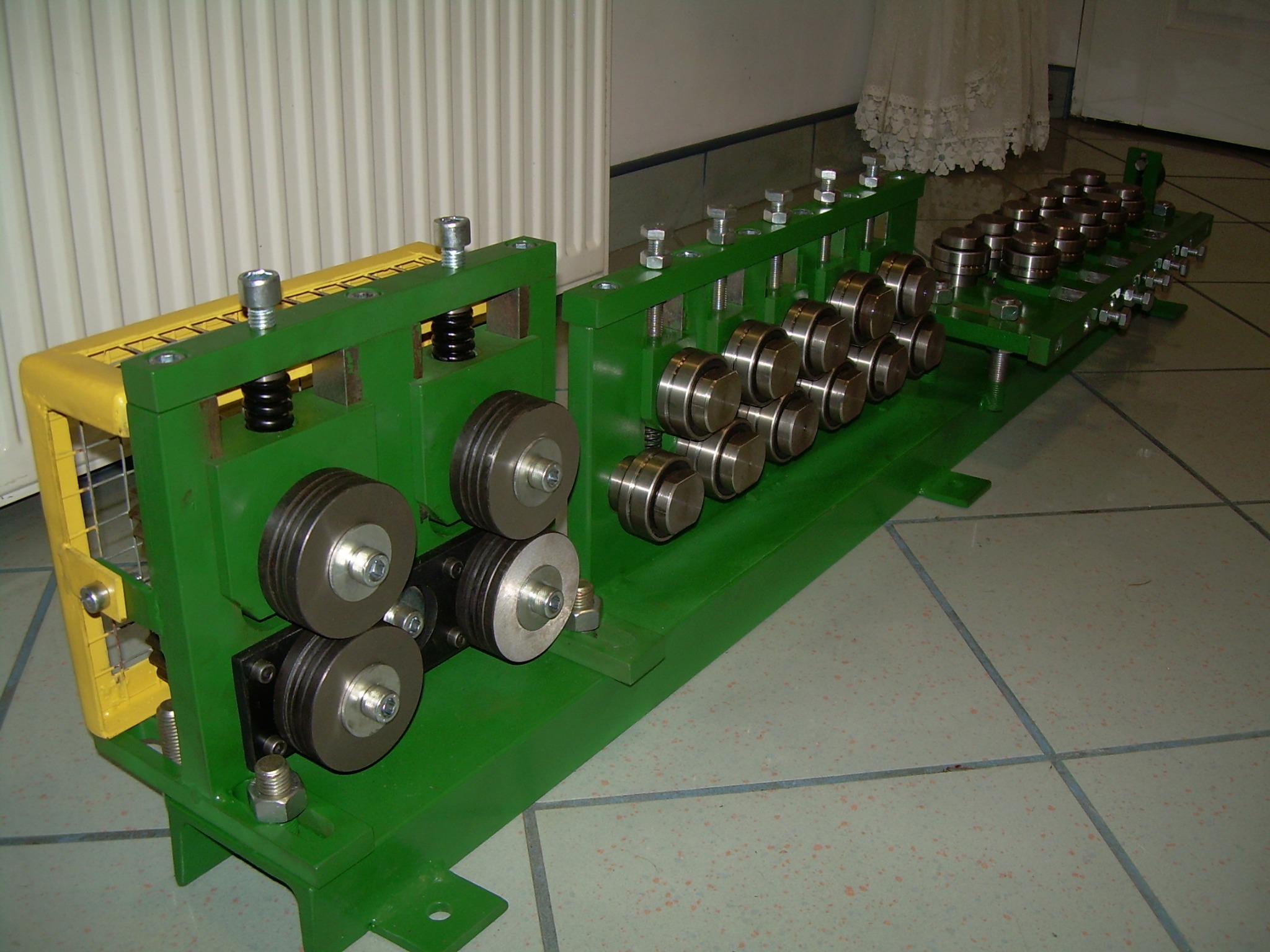 Zielona maszyna do obróbki metalu z szeregiem metalowych rolek i żółtą osłoną, ustawiona na kafelkowanej podłodze, z widocznym grzejnikiem w tle.