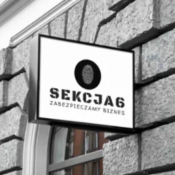 Szyld firmy ochroniarskiej 'SEKCJA6' z odciskiem palca na szarym, kamiennym murze budynku.