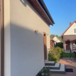 Elewacja budynku po remoncie z nowymi rynnami, kostką brukową prowadzącą do wejścia i rabatą kwiatową u podstawy ściany.