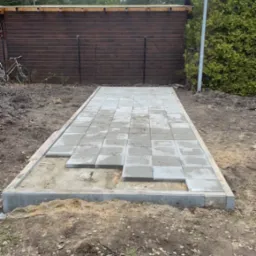 Przygotowanie podłoża pod ułożenie szarej kostki brukowej, widoczne obramowanie i częściowo ułożona nawierzchnia na piaszczystym podłożu, tło stanowi drewniany budynek gospodarczy i zieleń.