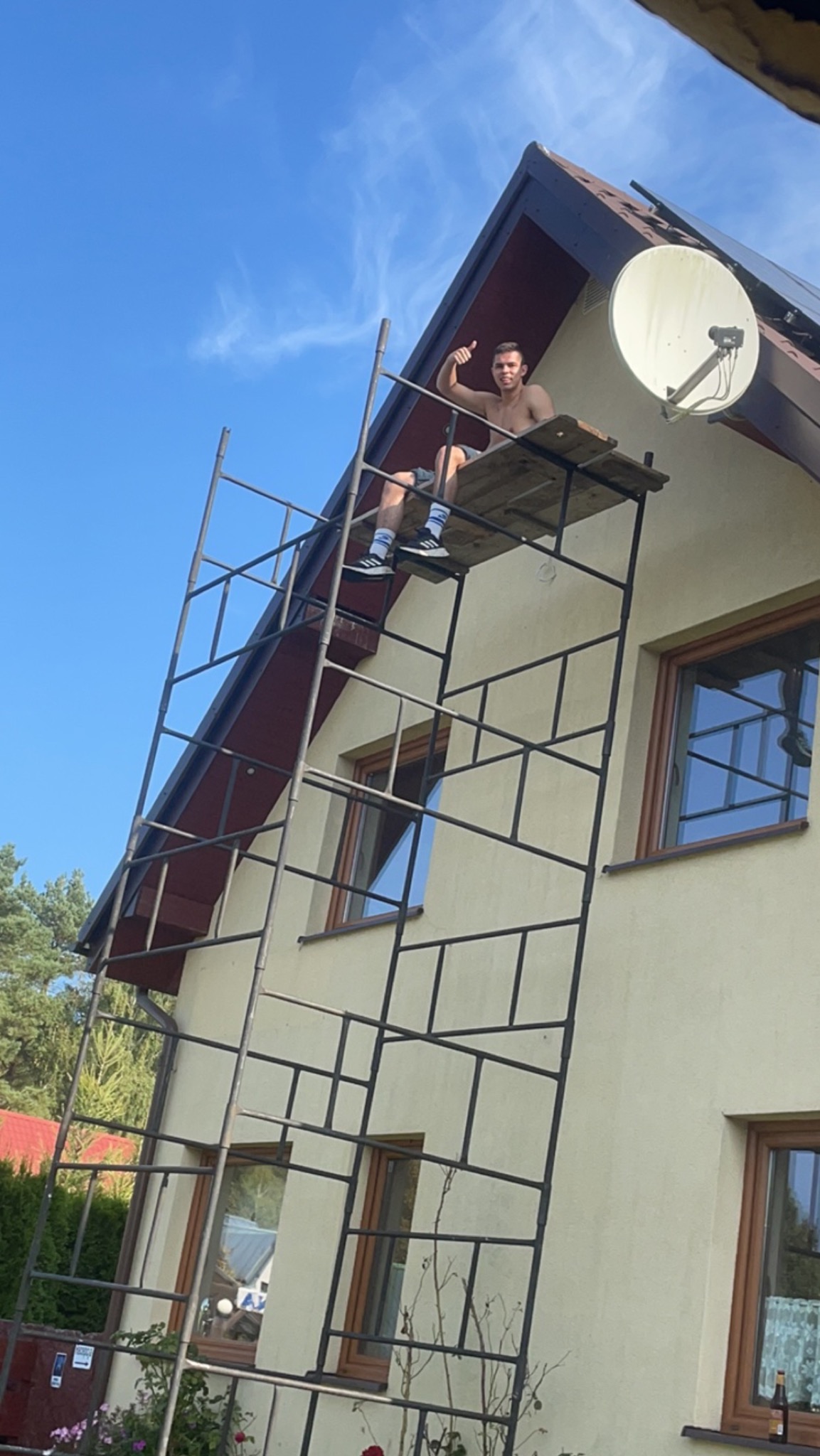 Mężczyzna na rusztowaniu przed domem, pozujący z uniesionym kciukiem, widoczna elewacja domu, okna z drewnianymi ramami, antena satelitarna i panele słoneczne na dachu.