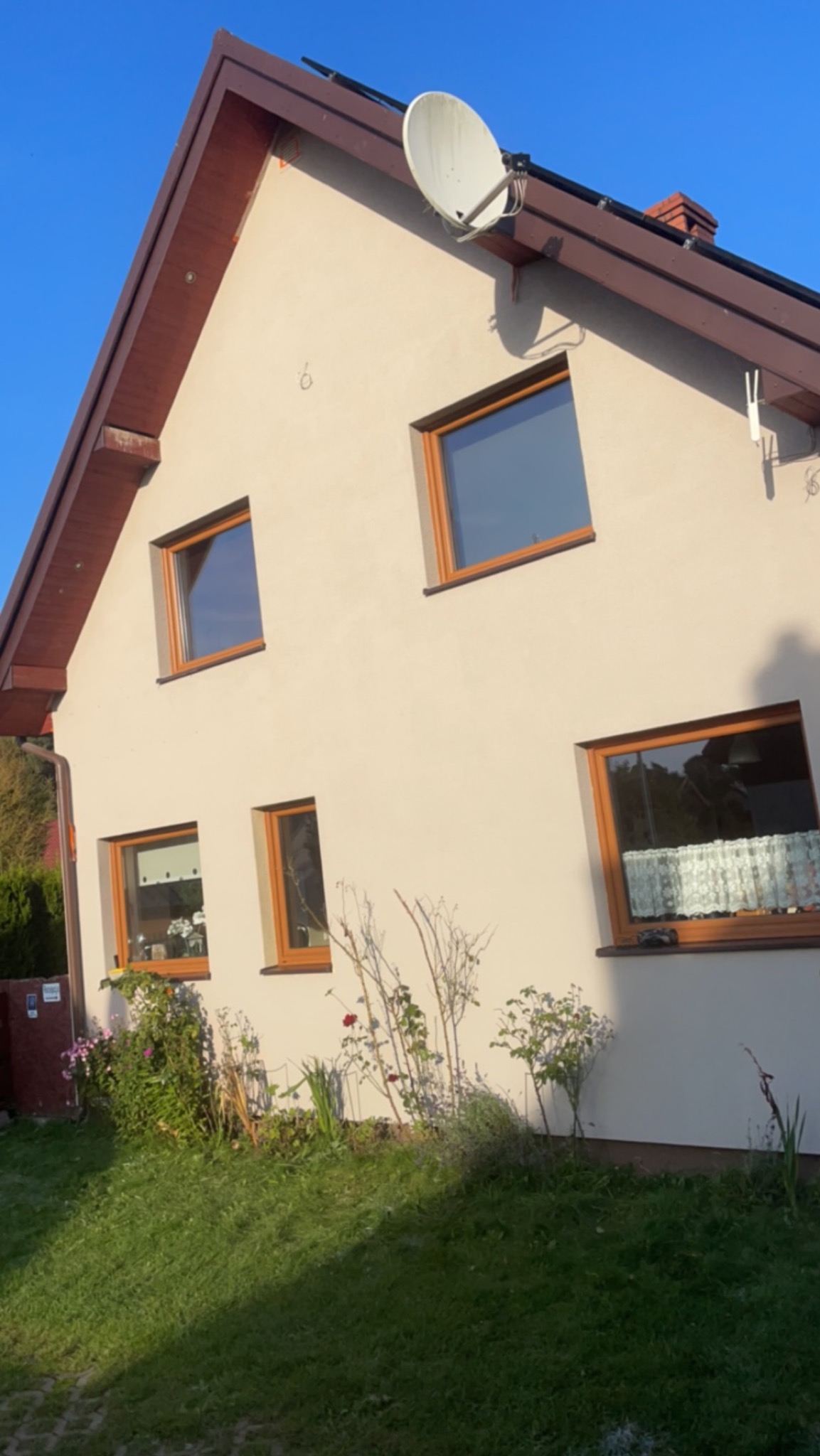 Jasny, otynkowany dom jednorodzinny z brązowymi ramami okiennymi i dachem, antena satelitarna na dachu, ogród z kwiatami u podstawy ściany.