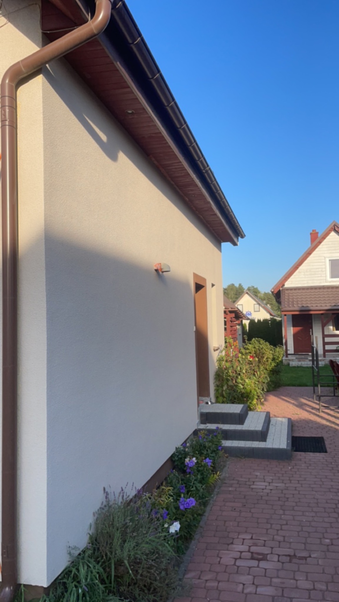 Elewacja budynku po remoncie z nowymi rynnami, kostką brukową prowadzącą do wejścia i rabatą kwiatową u podstawy ściany.