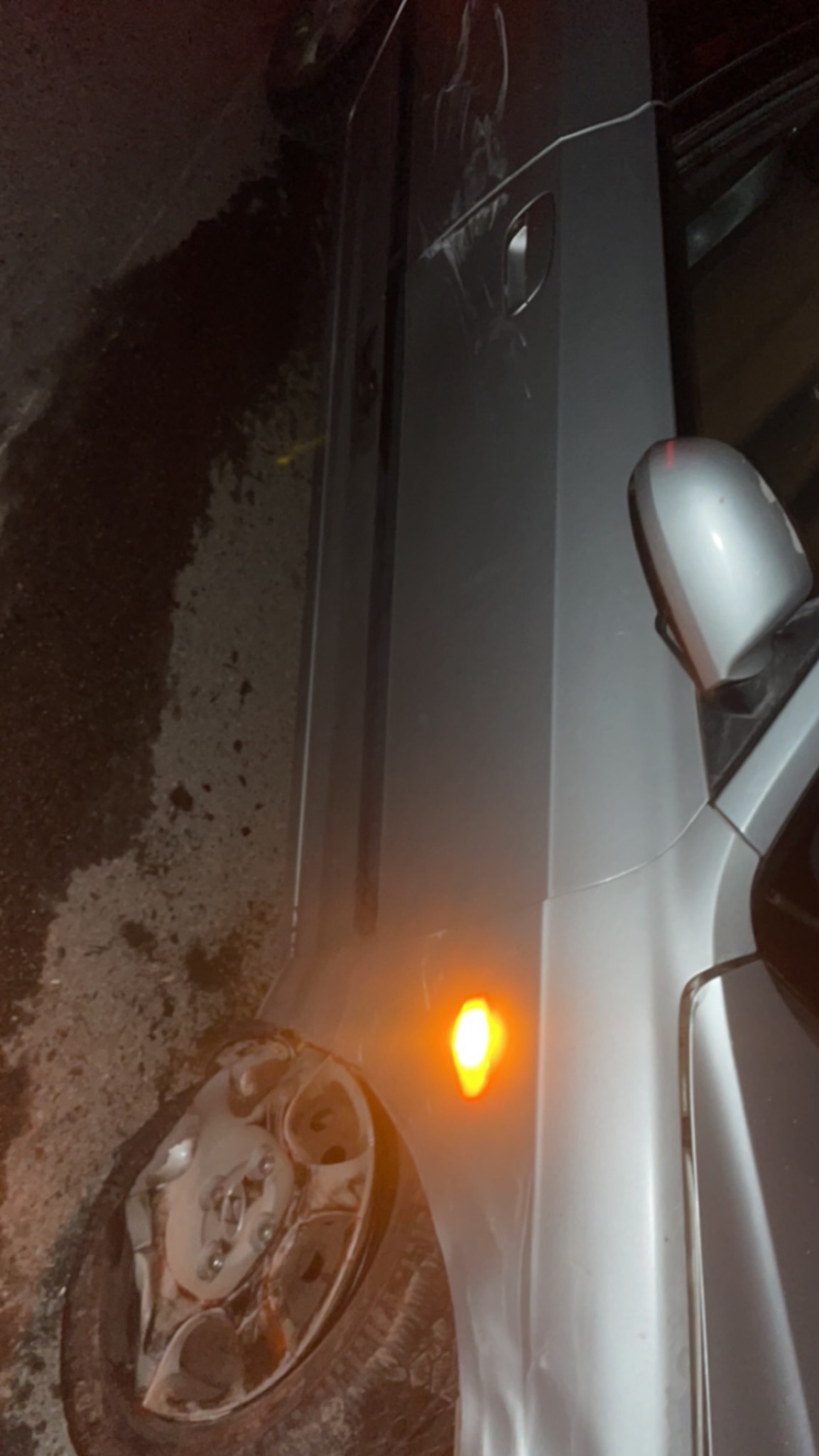 Uszkodzony srebrny samochód z włączonym kierunkowskazem, widoczne zarysowania na karoserii i zniszczona opona na tle ciemnego podłoża.