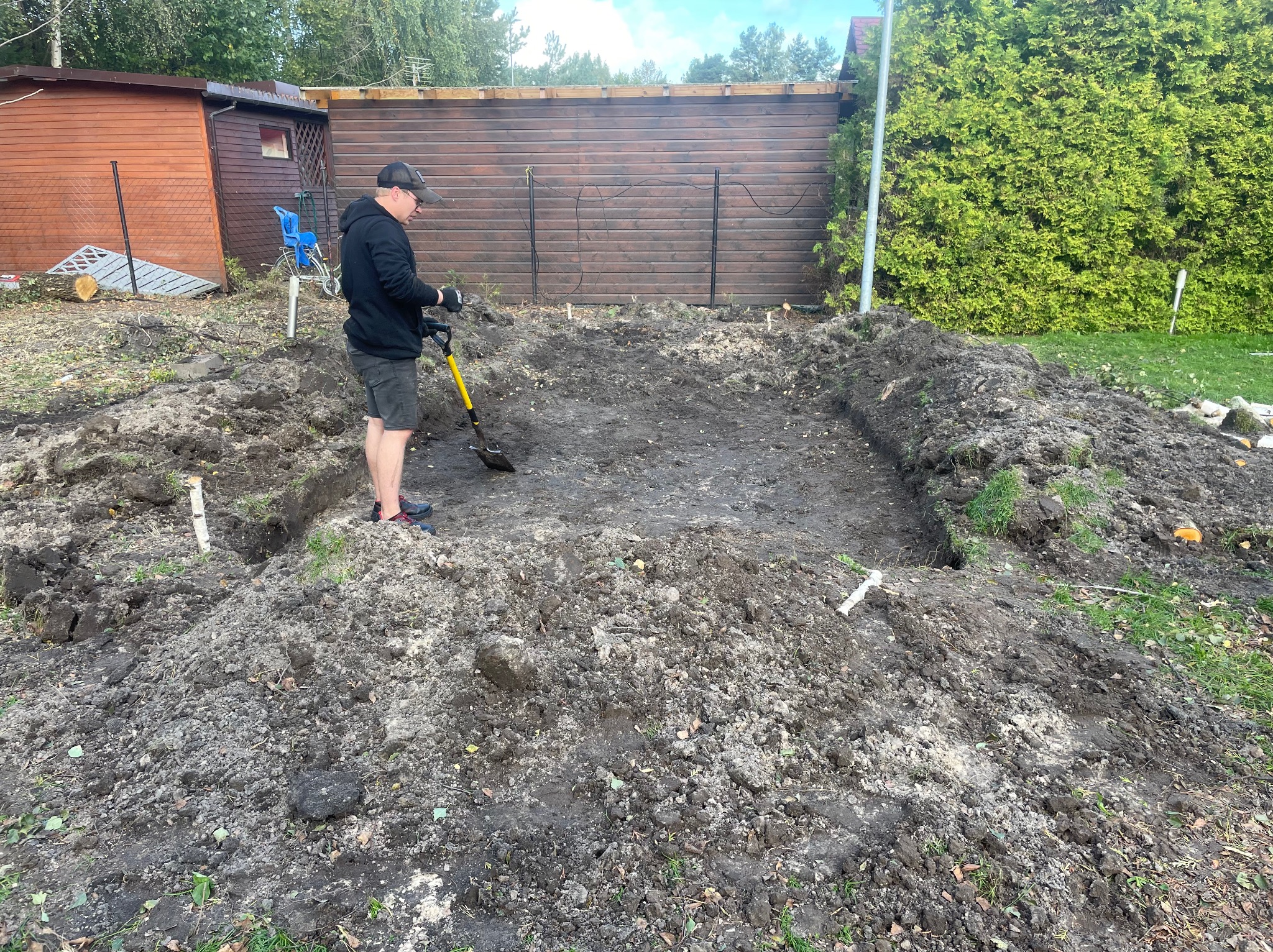 Mężczyzna w czarnej bluzie kopie dół pod staw szpadlem z żółtą rączką na tle drewnianego płotu i zielonych krzewów.