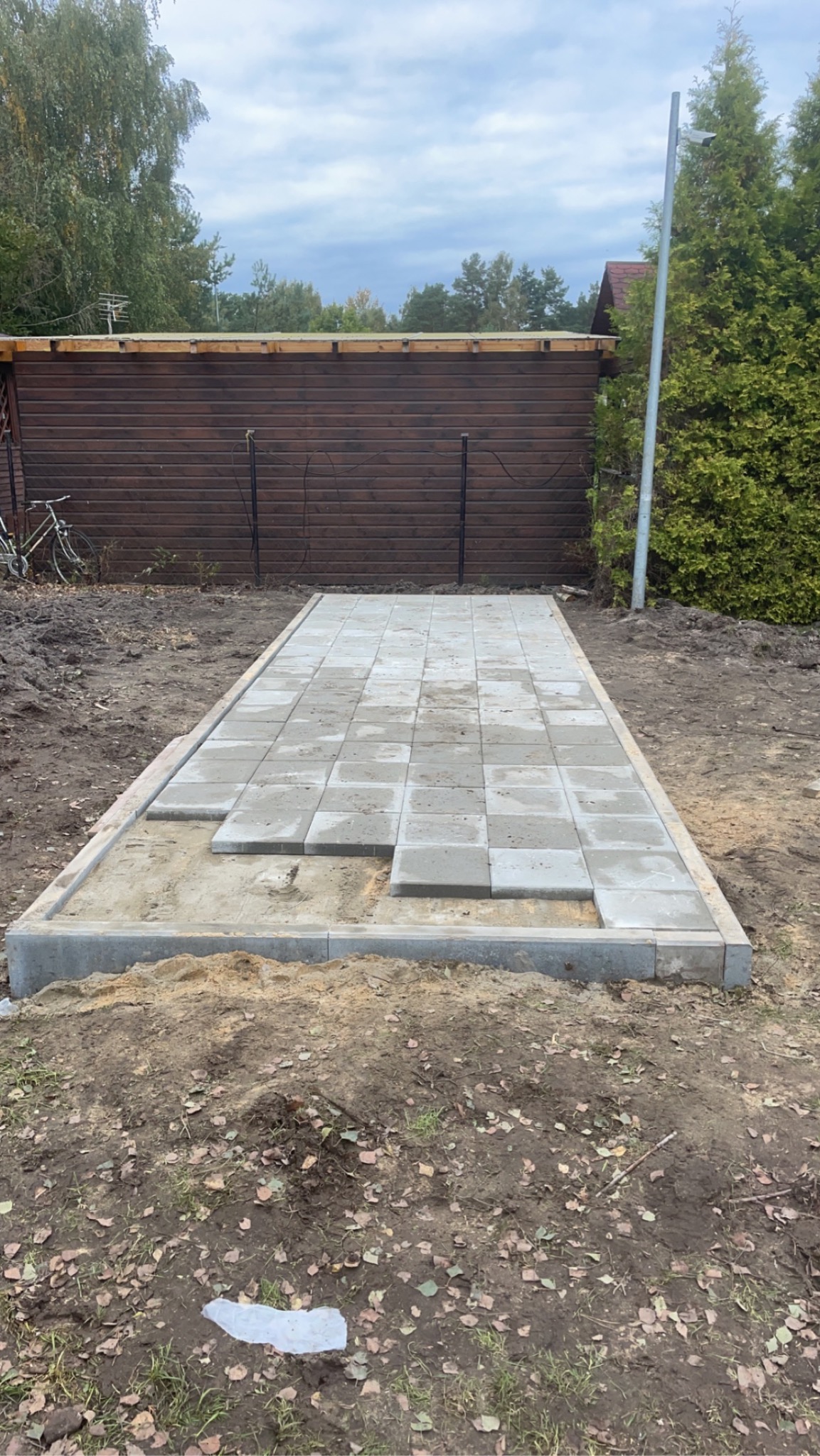 Przygotowanie podłoża pod ułożenie szarej kostki brukowej, widoczne obramowanie i częściowo ułożona nawierzchnia na piaszczystym podłożu, tło stanowi drewniany budynek gospodarczy i zieleń.