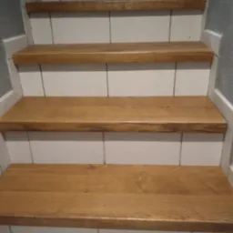 Fragment schodów z drewnianymi stopniami o ciepłym odcieniu, osadzonymi na białych, kwadratowych płytkach, widoczne z dołu