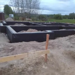 Zabezpieczony hydroizolacją fundament domu w trakcie budowy, widoczne drewniane stemple i szalunki, w tle zielona łąka i pochmurne niebo.