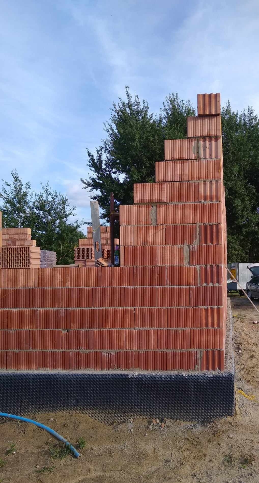 Wznoszenie ściany z czerwonych bloczków ceramicznych na fundamencie z hydroizolacją, widoczne narzędzia i poziomica, w tle drzewa i niebo.