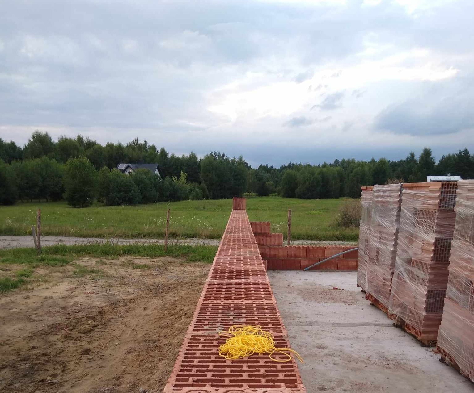 Wstępny etap budowy domu z cegły ceramicznej, widoczne ułożone cegły na fundamencie oraz zapas cegieł na paletach, w tle zielona łąka i las.