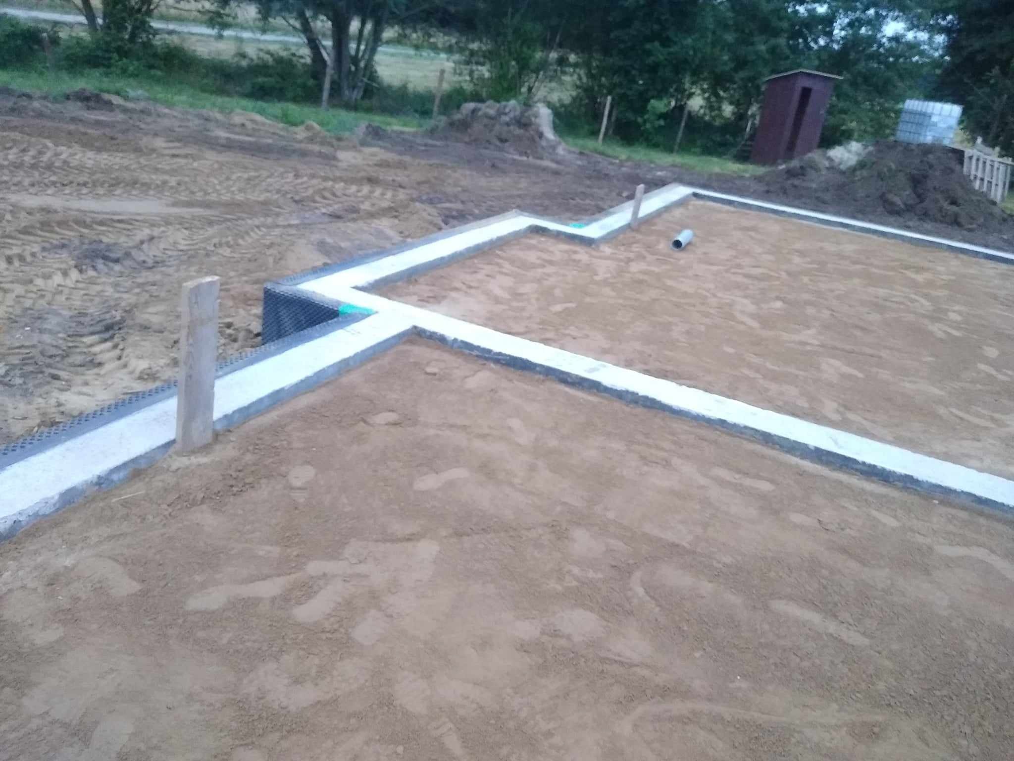 Wylane fundamenty pod budynek, widoczna izolacja pionowa i elementy kanalizacji, w tle teren budowy z barakiem i stertą ziemi.