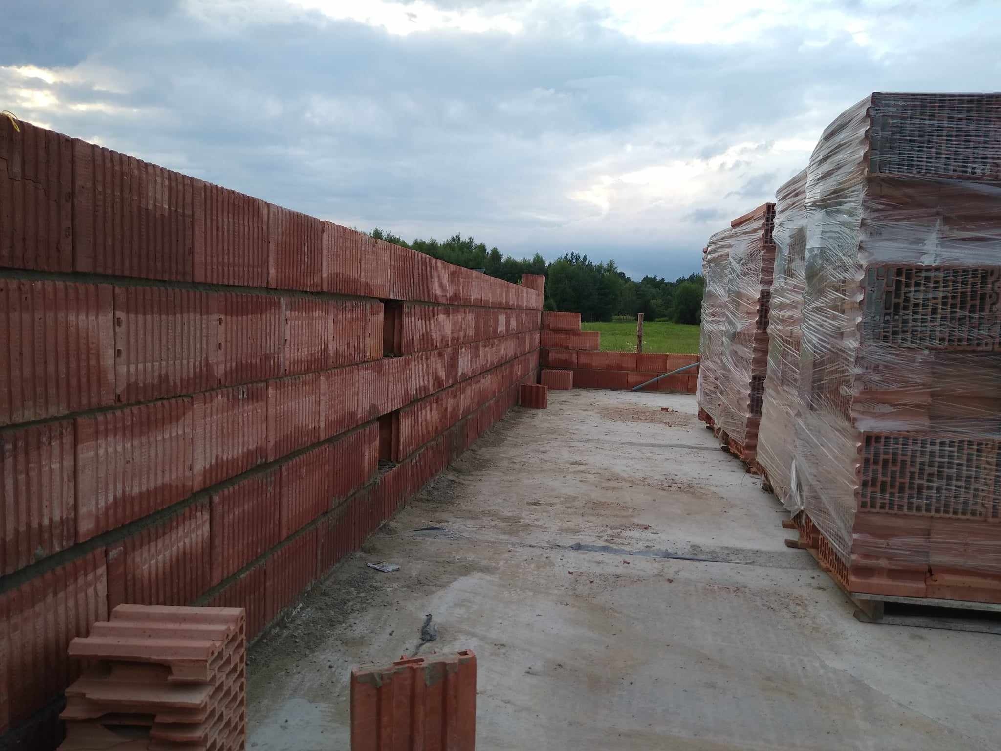 Wznoszenie ściany z czerwonych, porowatych pustaków ceramicznych na budowie domu, widoczne zapasy materiału na paletach, pochmurne niebo w tle.