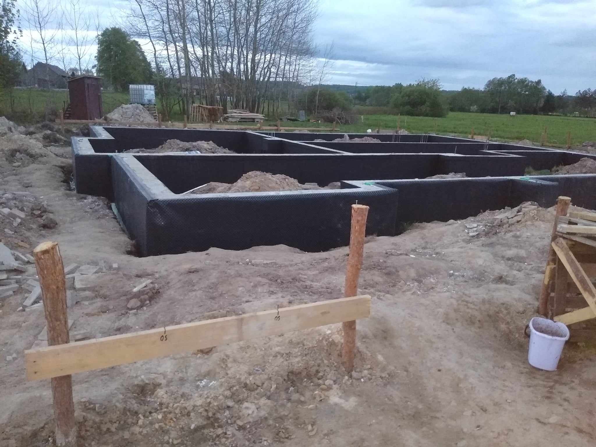 Zabezpieczony hydroizolacją fundament domu w trakcie budowy, widoczne drewniane stemple i szalunki, w tle zielona łąka i pochmurne niebo.