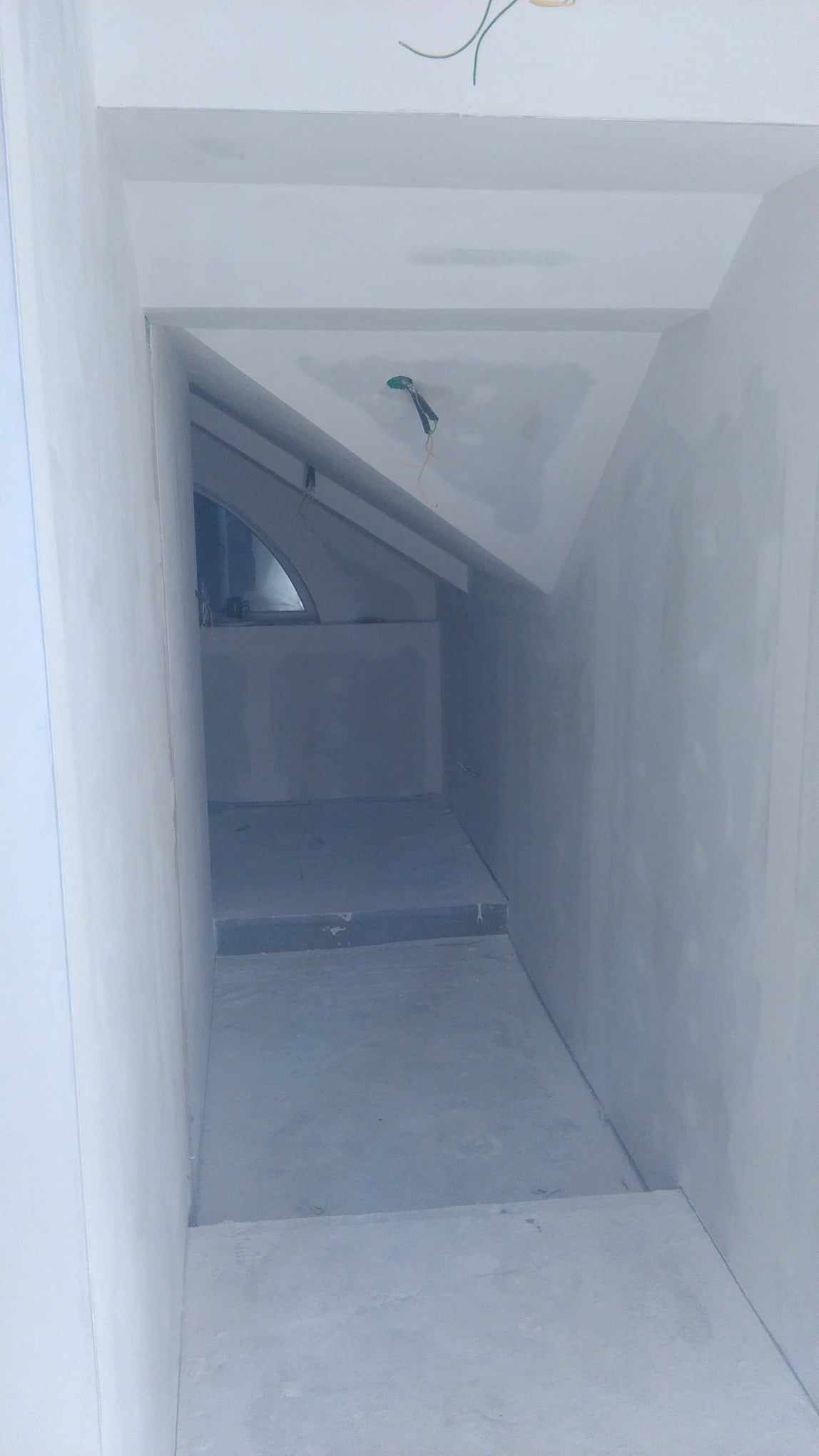 Wnętrze w trakcie remontu: surowe ściany i schody przygotowane do malowania, widoczne kable elektryczne wystające z sufitu, małe okno w tle.