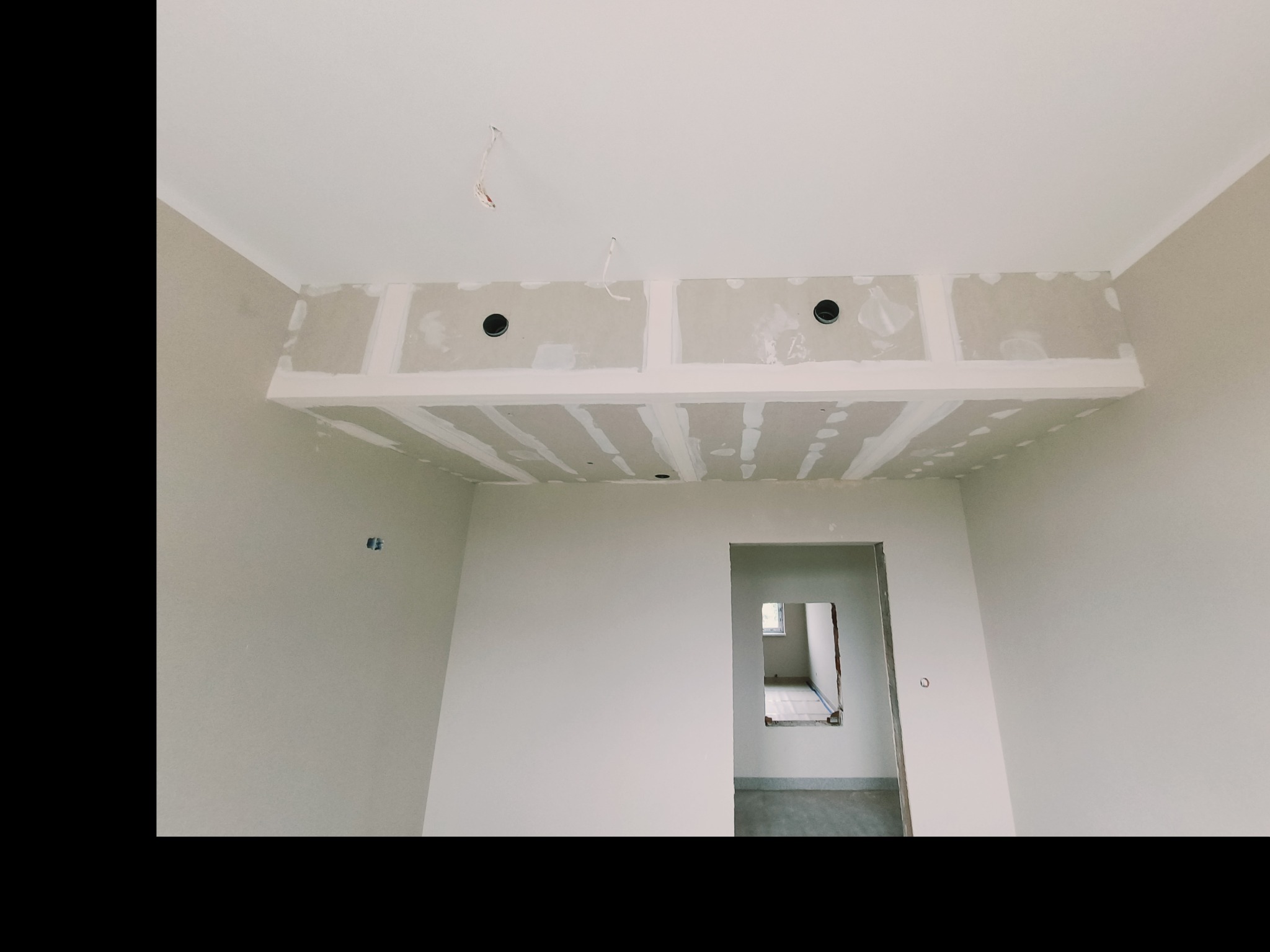 Wnętrze w trakcie remontu: sufit podwieszany z płyt karton-gips, ściany w jasnym odcieniu, widoczne otwory na oświetlenie i fragment instalacji elektrycznej.